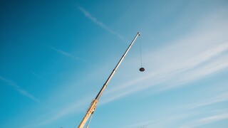 A long crane arm extends into a vibrant blue sky, suspending a dark, spherical object. The scene is simple, yet striking, highlighting the cranes reach and the weight it carries.
