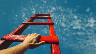 A male hand holds onto the crossbar of a red wooden staircase leading to the blue sky